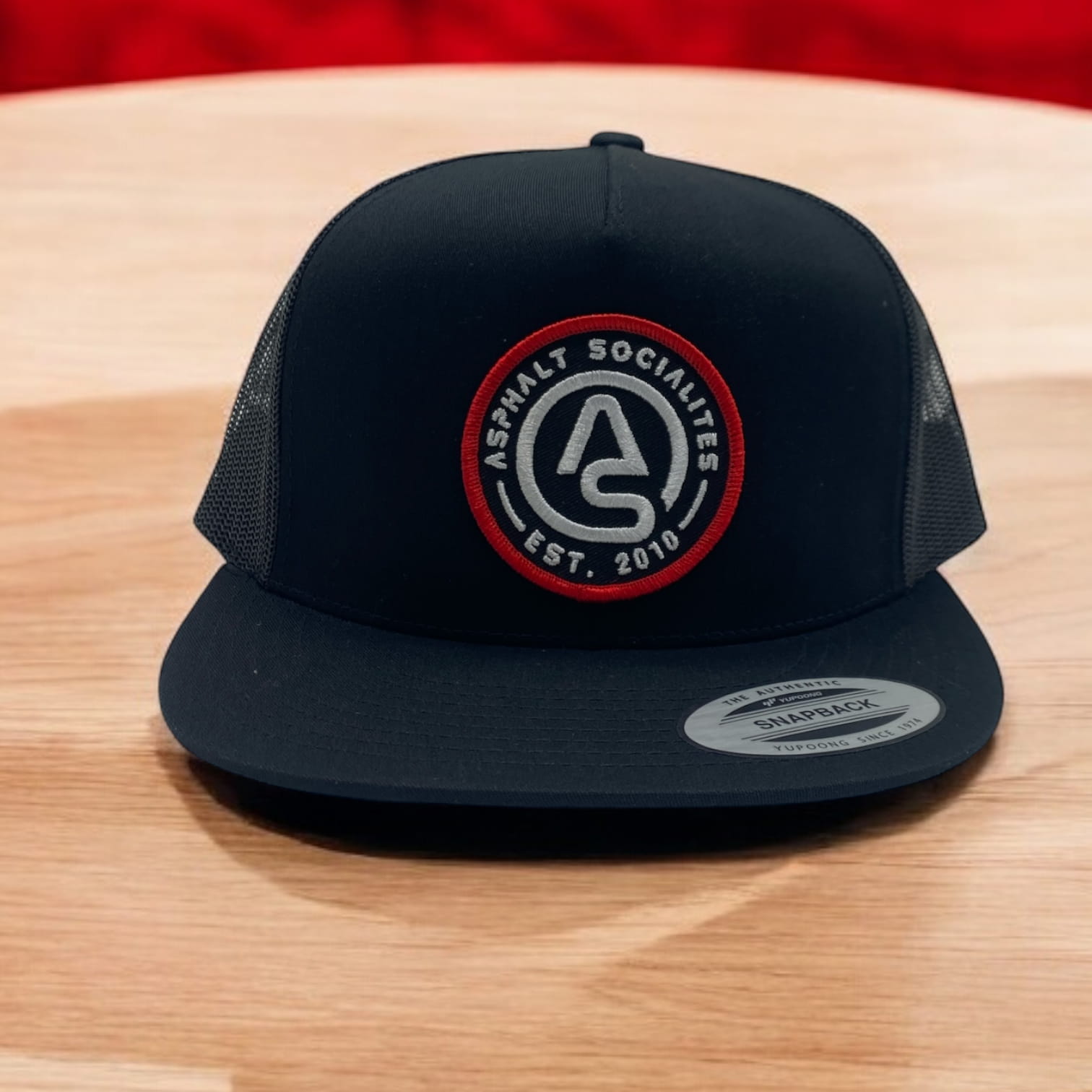 Black Yupoong snapback hat with Asphalt Socialites round patch logo and Est. 2010 text in red, gray, and white.