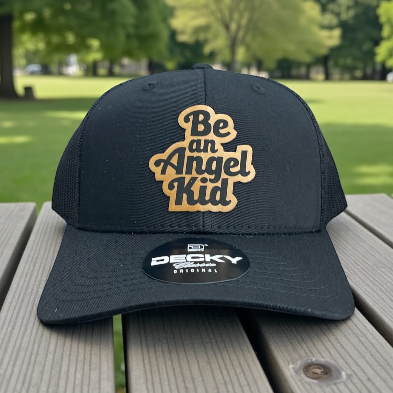 Black Decky trucker hat with tan Be an Angel Kid logo patch on the front, mesh back, curved bill.