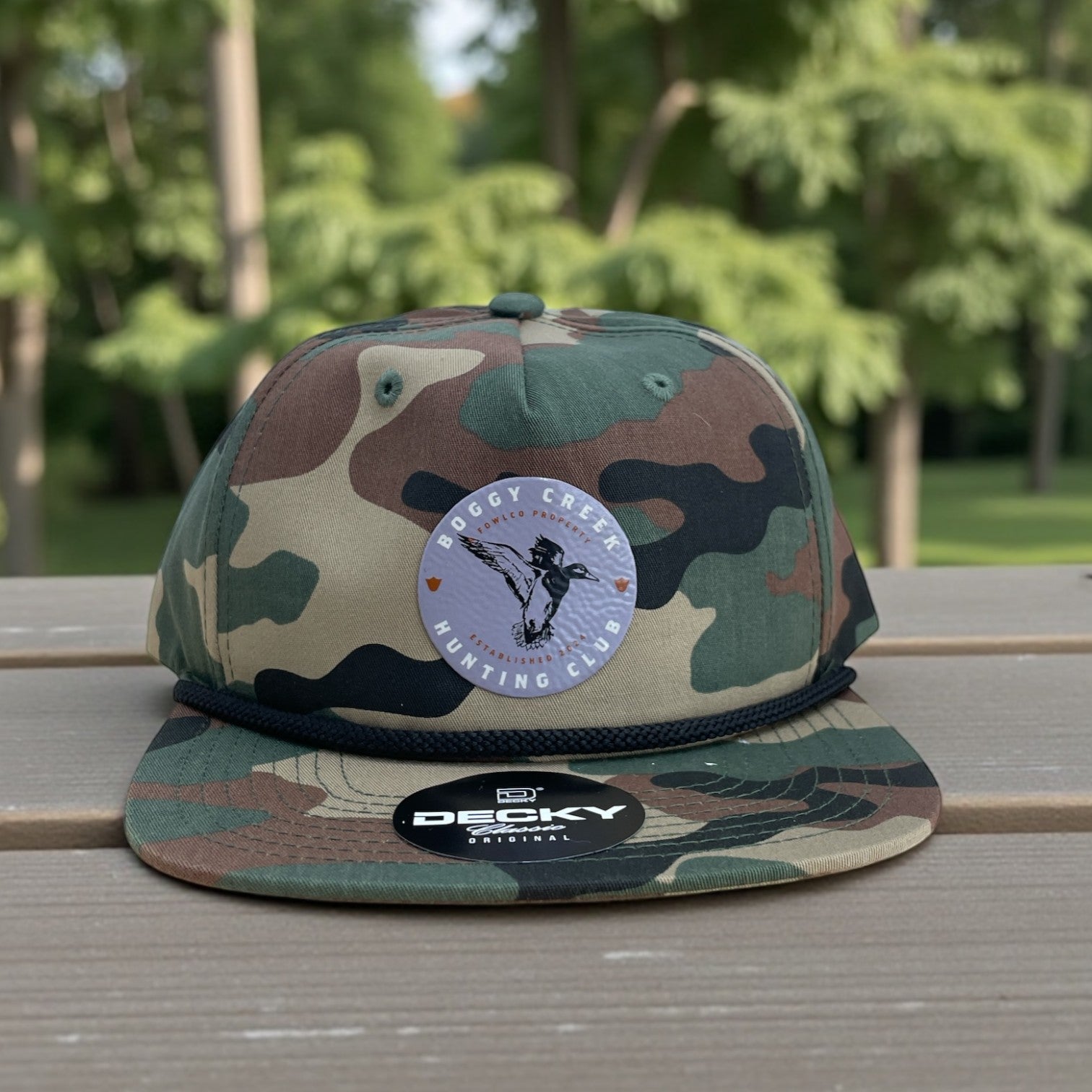 Camo flat-bill cap with Boggy Creek Hunting Club round patch and black rope detail on the crown.