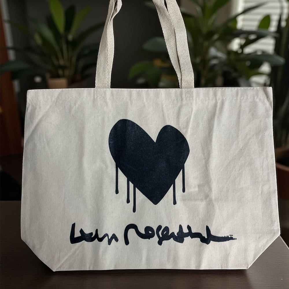 Natural canvas tote bag with black dripping heart graphic and handwritten-style logo, held by hand indoors.