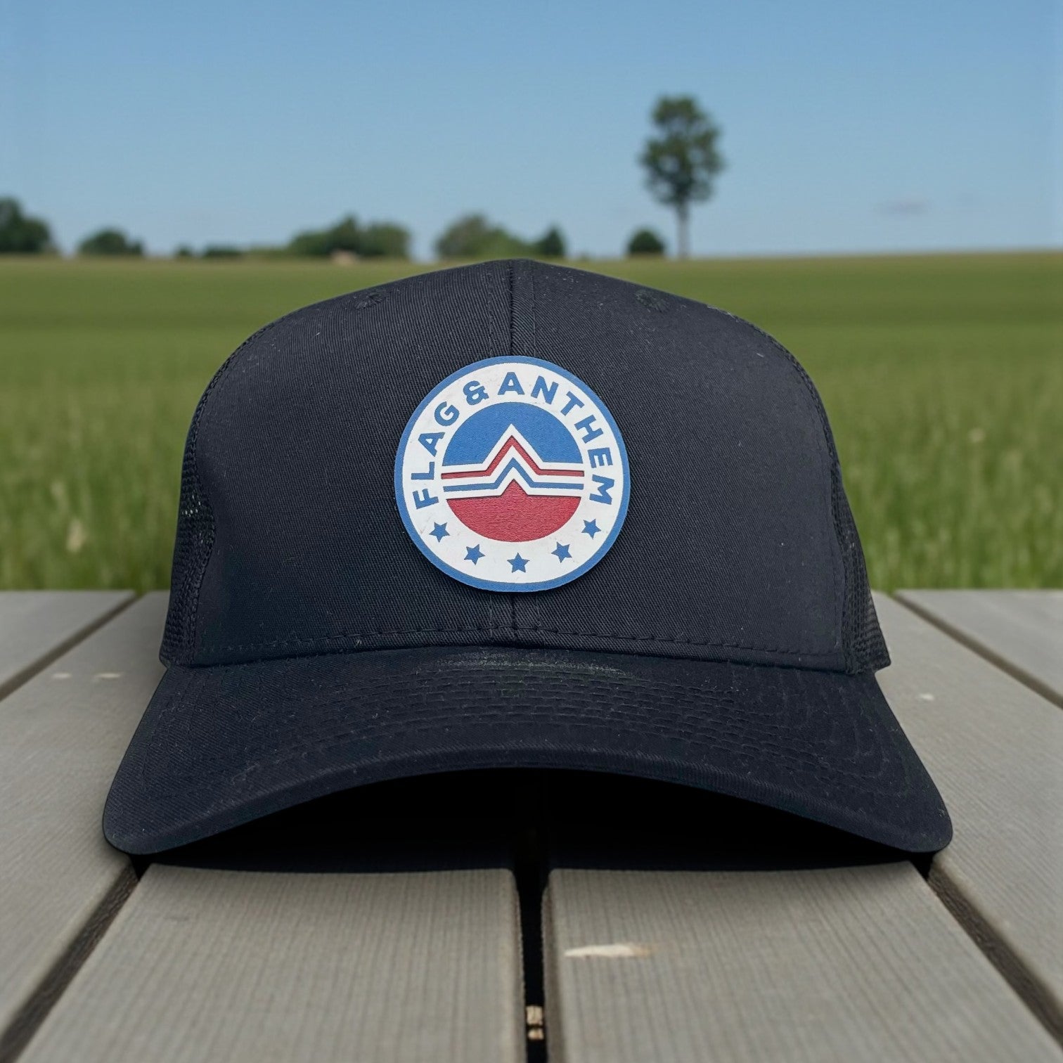 Black mesh-back trucker hat with Flag & Anthem round patch logo, shown on outdoor picnic table.