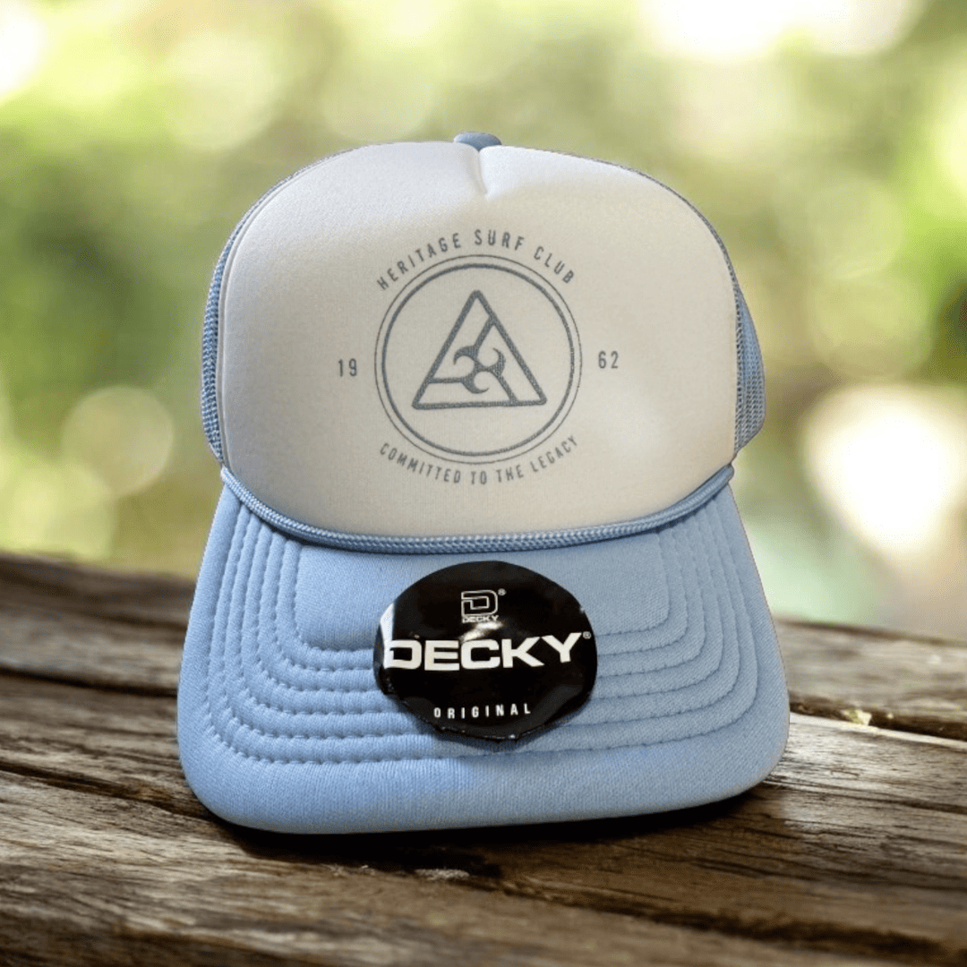 Light blue and white Heritage Surf Club trucker hat with 1962 badge logo, photographed on wooden table.