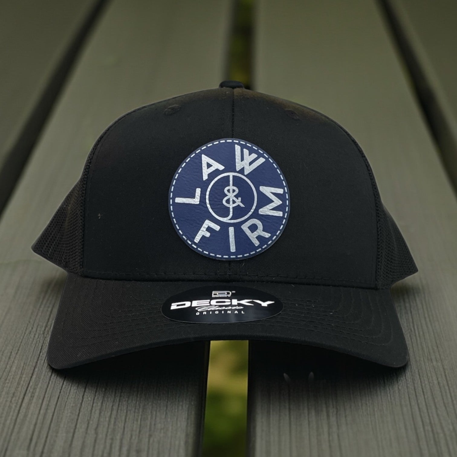 Black trucker hat with blue round LAW & FIRM logo patch on the front, photographed on a picnic table.