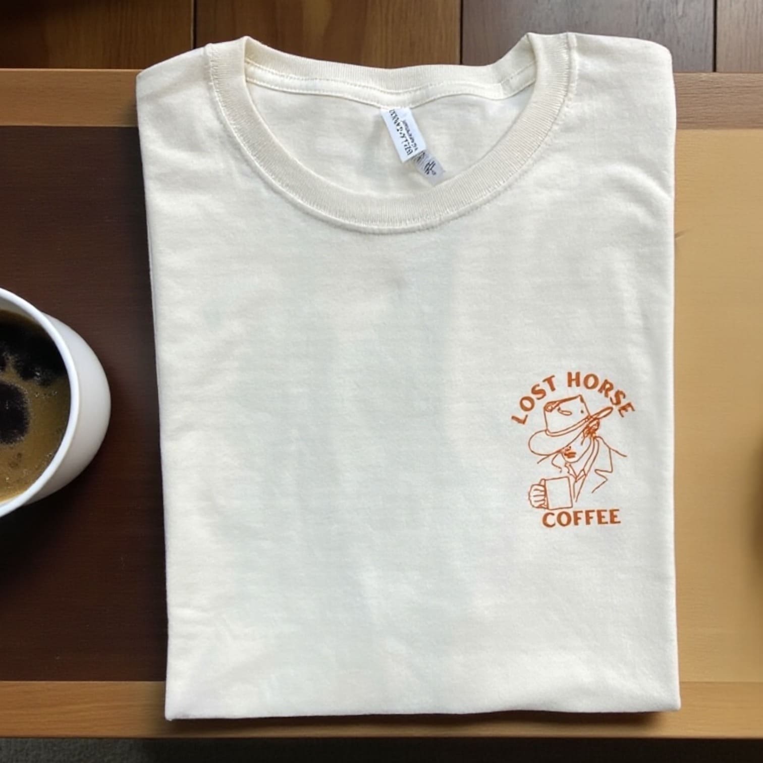 Cream Lost Horse Coffee t-shirt folded flat with small cowboy logo on chest, next to coffee cup.