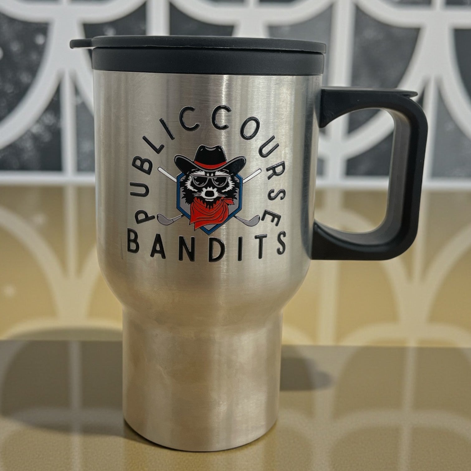 Stainless steel travel mug with handle and lid featuring Public Course Bandits logo with masked raccoon graphic.