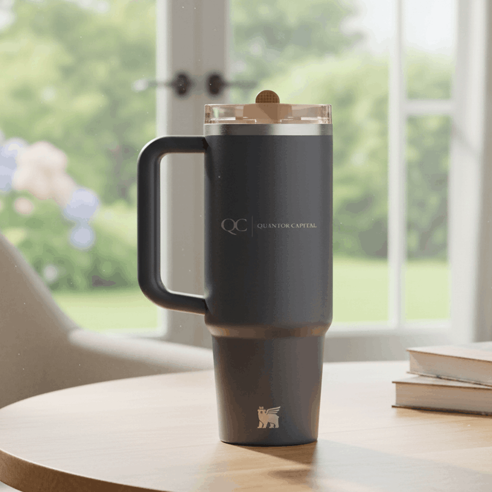 Black handled travel tumbler with QC Quantor Capital logo, clear lid, on round table by window in bright room.