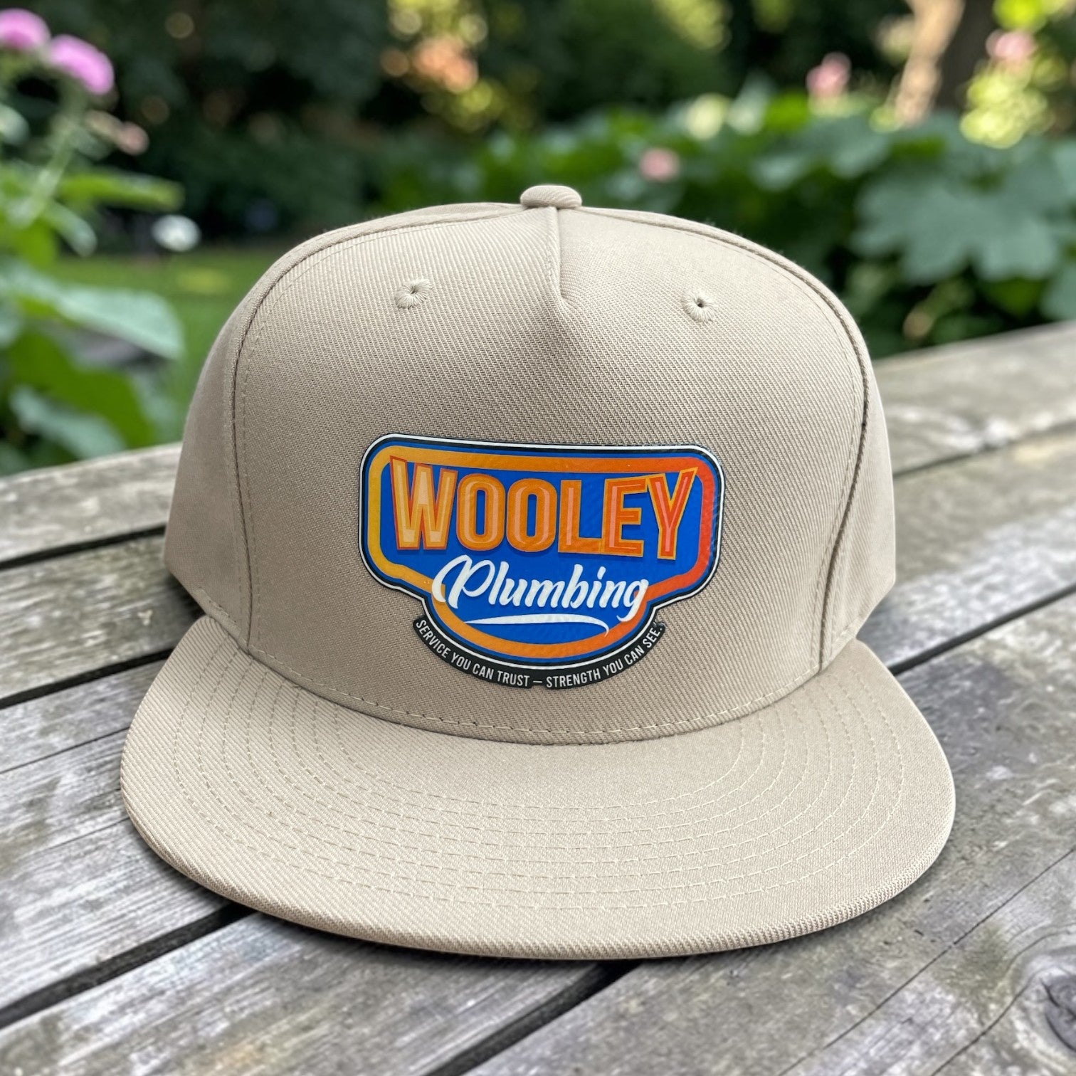 Beige flat-bill snapback hat with Woolley Plumbing logo patch on the front panel, outdoors on wood table.